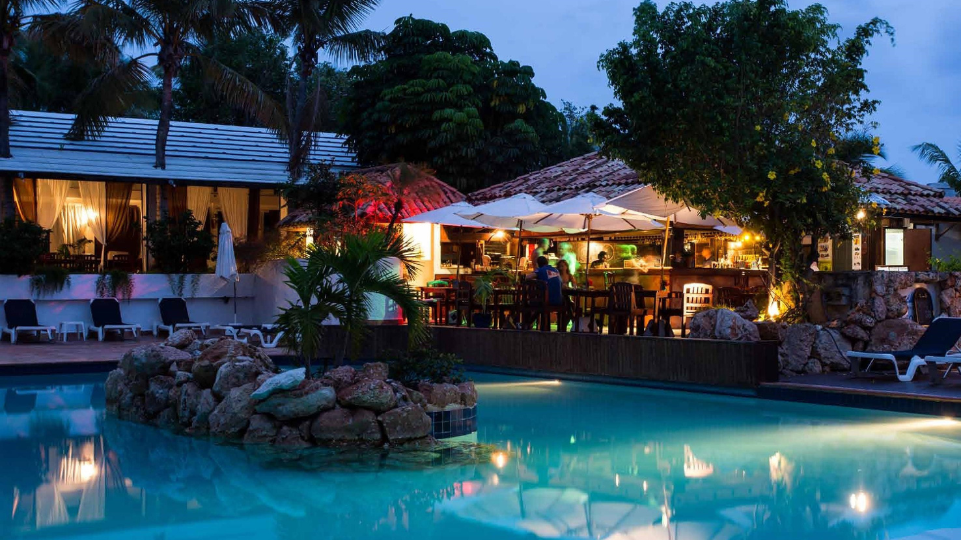 Night time view of rocky island with palm tree and restaurant alongside pool of Sapphire Beach Club Resort in Cupecoy